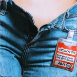 Close-up of a blue denim jacket with a red event badge clipped to the chest, showing the word 'SCOOTER' and small print badges.