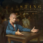 Man with a blue shirt sits at a wooden table, arms outstretched, in front of a dark poster that says 'STING The Night Watch Live at the Rijksmuseum'