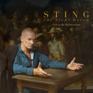 Man with a blue shirt sits at a wooden table, arms outstretched, in front of a dark poster that says 'STING The Night Watch Live at the Rijksmuseum'