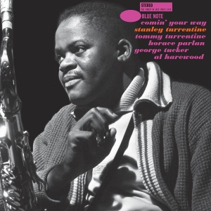 Black-and-white close-up of a jazz saxophonist on stage, instrument visible on the left and a cozy sweater collar seen in the foreground.
